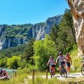 A vibrant outdoor scene in the Ardeche region showcasing a stunning landscape with cliffs A vibrant outdoor scene in the Ardèche region, showcasing a stunning landscape with cliffs, lush green forests, and the mesmerizing Ardèche River. In the foreground, a group of diverse individuals, dressed in modest sports attire, engage in activities like canoeing and rock climbing, exuding a sense of adventure and camaraderie. The middle ground features cyclists on mountain bikes navigating a scenic trail, while a nearby cave entrance hints at exciting spelunking opportunities. The background showcases dramatic limestone cliffs against a clear blue sky, with sunlight filtering through the trees, creating a warm and inviting atmosphere. The image should be realistic with bright, natural lighting, captured from a slightly elevated angle to encompass the beauty and energy of outdoor sports in Ardèche.